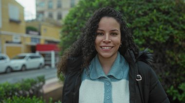 Parktaki İspanyol kadın ceketini giyiyor ve yeşillik yakınında gülümsüyor ve bulutlu bir günde açık havada canlı şehir hayatını gösteriyor..