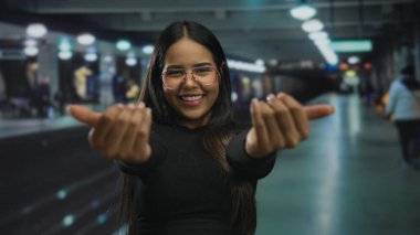 Latin bir kadın, kapalı bir tren istasyonunda neşeli bir şekilde gülümsüyor. Tren peronunun yanında kendine sarılırken sıcaklığını ve neşe saçıyor..