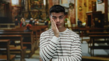 Çizgili gömlekli genç bir adam kilisenin içinde elleri yüzünde düşünceli görünüyor, huzurlu bir atmosferde boş sıralarla çevrili..
