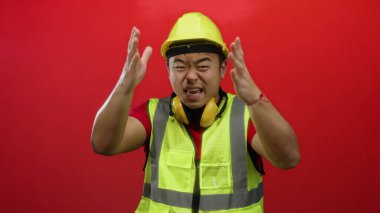 Sarı güvenlik yeleği giyen ve kasketli genç adam canlı kırmızı bir arka planda duruyor. İnşaata hazırlık ve profesyonelliği somutlaştırıyor..