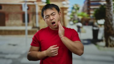 Kırmızı tişörtlü genç adam, şehir arka planında bulanık bir diş ağrısı çekiyor. İfadesinde acıdan rahatsız olduğunu ifade ediyor..
