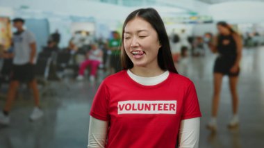Havaalanı terminalinde gülümseyen bir kadın. Arka planda bulanık gezginlerle gönüllü tişörtü giyiyor. Kapalı alanda canlı bir atmosfer yakalıyor..