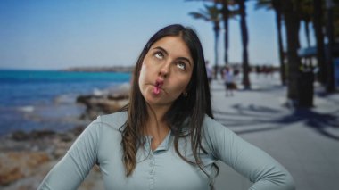 Açık mavi gökyüzünün altında palmiye ağaçlarıyla deniz kenarında neşe içinde poz veren kadın..