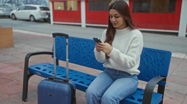 Sokakta bir bankta oturan bir kadın mavi bavullu bir akıllı telefon kullanıyor, şehir içi bir seyahat sahnesini kaldırımda günlük kıyafetleriyle çekiyor..