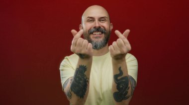 Kel, sakallı, parlak kırmızı bir duvara karşı kalp hareketi yapan, sıradan, dostane bir ifade sergileyen, kel ve dövmeli bir adam..