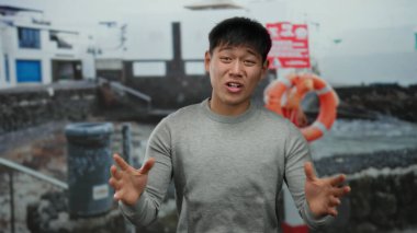 Gri kazaklı genç adam sahilde duygularını ifade ediyor. Arka planda can yelekleri ve binalar var. Sahilde bir açık hava sahnesi sergiliyor..