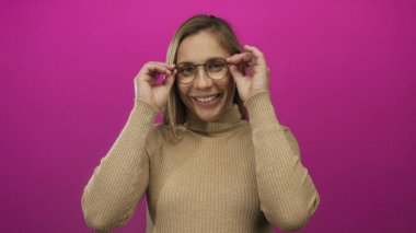 Boğazlı kazakla gülümseyen, canlı pembe arka planda duran, neşeli ifadeli gözlükleri ayarlayan ve neşeli havayı yansıtan rahat duruşu olan kadın..