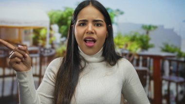 Kadın, restoranın içindeki bir sunucuyu işaret ederken parmağını kaldırıyor. Genç Latin kökenli bir kadın terasta dikiliyor ve kızgın görünüyor..