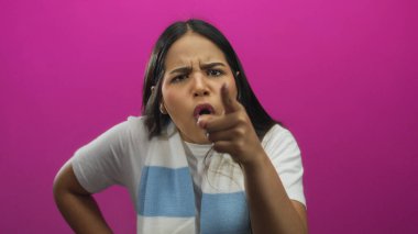 Parmağını kaldırmış bir kadın stüdyo ortamında kızgın görünüyor. Pembe arka plan duvarında Latin kökenli genç bir kadının ifadesini yakalıyor..