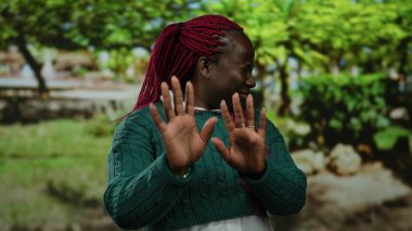 Kırmızı örgülü Afro-Amerikalı bir kadın parkta yeşil bir kazak giyip doğal ortamda duygularını ifade ederek reddediyor..