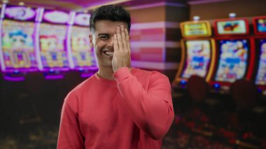 Canlı bir kumarhanede genç bir adam renkli kumar makinelerinin yanında neşeli bir ifade ve rahat bir atmosfer sergileyerek durur..