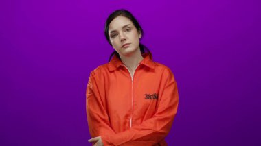 Turuncu hapishane tulumlu kadın canlı mor bir arka plana karşı duruyor, esmer saçlar yüzünü çerçeveliyor, güçlü bir hapsetme ve tecrit havasını taşıyor..