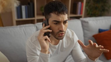 Genç bir adam telefonda konuşuyor. Oturma odasında ciddi bir ifade takınıyor, günlük giysiler giyiyor ve sıcak bir dekorla çevrili rahat bir koltukta oturuyor..