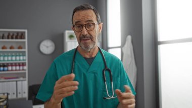 Önlük giyen ve steteskop kullanan İspanyol olgun bir adam hastane odasında duruyor. Sağlık hizmetlerini gösteren arka planda raflarla ve saatlerle tıbbi detayları anlatıyor..