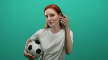 Futbol topunu tuhaf bir ifadeyle tutan kadın yeşil bir zemine karşı duruyor. Günlük beyaz bir gömlek giyiyor. Düşünceli ve enerjik bir görünümü var..