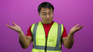 Emniyet yeleği giyen genç adam avuçlarını pembe arka plana dayamış gülümsüyor.