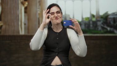 Kadın, güneşli açık hava ortamının altında antik Roma harabelerinin önünde gururla kredi kartını gösterirken iyi bir jest yapıyor..