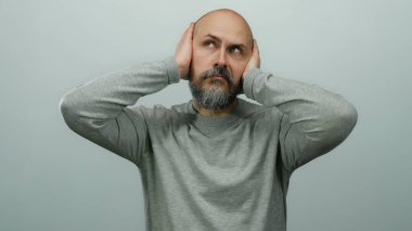 Kulakları elleriyle kapatan, beyaz arka planda hayal kırıklığını ifade eden, günlük gri süveter giyen bir adam..