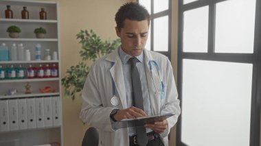 Klinik üniformalı, steteskop ve not panosuyla genç İspanyol doktor hastane odasındaki iş yerinde profesyonelliği ve bağlılığı yansıtıyor..