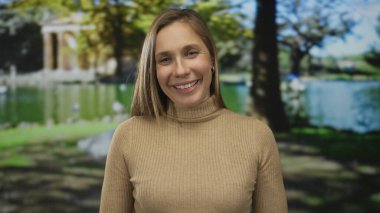 Sarışın bir kadın parkta gülümsüyor. Etrafı gür ağaçlar ve dingin bir göl ile çevrili. Sıcak ve davetkar bir atmosfer yaratıyor..