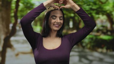 Parkta taç giyen bir kadın elleriyle kalp hareketi yapıyor, neşe ve kendini beğenmişlik gösteriyor, gür ağaçlarla çevrili, açık havada zarafet ve oyunculuğu somutlaştırıyor.