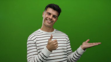 Genç adam neşeli bir şekilde göz kırpan bir ifadeyle izole edilmiş, çizgili bir süveterle yeşil bir arka plana işaret ediyor..