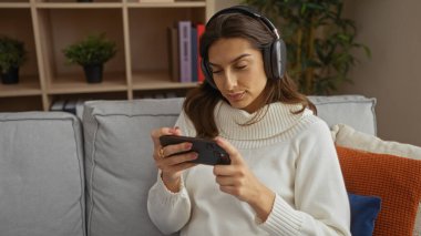 Oturma odasında kulaklıkla akıllı telefon oyunu oynayan bir kadın, koltukta minderlerle ve bitkilerle oturuyor, rahat bir yaşam tarzı sergiliyor..