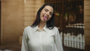 Sıcak bir arka planda, neşeli ve kaygısız bir ruh hali içinde, enerjik bir sokakta dikilmiş, dili dışarı doğru gülümseyen bir kadın..