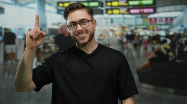 Sakallı, gülümseyen ve yukarıyı gösteren genç bir adam kalabalık bir havaalanı terminalinde duruyor. Arka planda insanlar var. Seyahat ve heyecan hissi taşıyor..