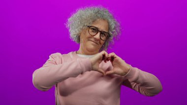 Kıvırcık saçlı, gözlüklü ve pembe kazaklı bir kadın izole edilmiş pembe arka plana karşı duruyor. Elleriyle kalp şekli oluşturuyor. Sıcaklık ve pozitiflik saçıyor..