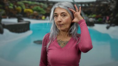 Gri saçlı yaşlı bir kadın oteldeki bir havuz kenarında dikkatle dinliyor, canlı bir açık hava atmosferinde merak ve bağlılık gösteriyor..