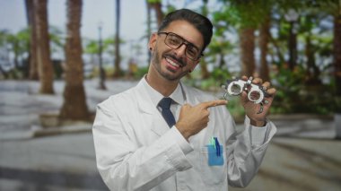 Gözlüklü ve beyaz önlüklü İspanyol adam gülümsüyor ve caddede phoropter lensini işaret ediyor..