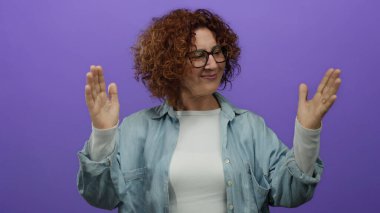 Orta yaşlı İspanyol bir kadın, elleri mor bir duvarın önünde, gözlük ve kot gömlek giyer ve büyük beden bir konsept taşır..