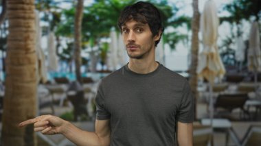 Gri gömlekli genç adam, havuz başında palmiye ağaçları ve kapalı şemsiyeli bir otel binasını işaret ediyor..