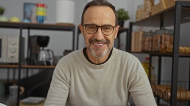 Kapalı bir ofis ortamında gülümseyen, raflar ve dekorasyon malzemelerinin yer aldığı rahat ve modern bir çalışma alanını sergileyen, profesyonel bir atmosferi ön plana çıkaran bir adam..