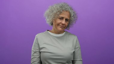 Gri saçlı bir kadın canlı mor bir arka plana karşı duruyor. Tarafsız bir gömlek giyiyor, çeşitli duygular ifade ediyor. Olgun güzellik ve kendine güven sergiliyor..