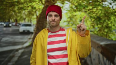Kırmızı bereli ve sarı ceketli genç adam şehir sokağında duruyor, baş parmağıyla aşağı işaret ediyor, arka planda canlı yeşil ağaçlar ve park edilmiş arabalarla çevrili..