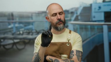 Kel sakallı, dövmeli ve siyah eldivenli bir adam deniz manzaralı bir yolcu gemisinde el kol hareketi yapıyor.