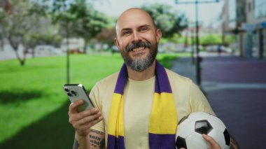 Parkta akıllı telefon ve futbol topu tutan adam sarı gömlek ve arka planda ağaçlar ve görünür dövmeler olan eşarp giyiyor.