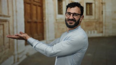 Eski bir kampüs binasının önünde elleri açık gülümseyen, gözlüklü ve açık mavi gömlekli İspanyol bir adam, bir üniversitede hoş geldin hissi uyandırıyor..