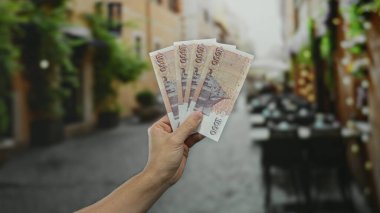 Şehir caddesindeki terasta ekonomik ve seyahat temalarını yakalayan, açık hava restoranlarını bulanık gösteren, İzlanda Krona banknotları tutan bir adam..