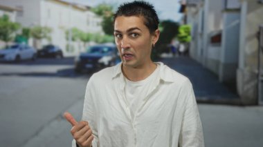 Beyaz tişörtlü genç İspanyol adam, güneşli şehir sokağında başparmağını yana gösteriyor; belirsizlik seçimi.