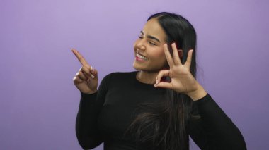 Gülümseyen bir kadın mor arka planda siyah uzun kollu bluz giyerek iyi bir hareket yapıyor..