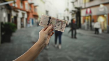Kalabalık bir şehir sokağında, arka planda bulanık insanlarla birlikte Suudi riyal banknotunu elinde tutarak, orta doğu para birimine odaklı bir kentsel çevre yakalıyor..