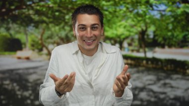 Açık havada güneşli bir parkta gülümseyen genç bir İspanyol yeşil ağaçlarla çevrili beyaz bir gömlek giyip sakin ve rahat bir atmosfer yaratıyor. Yaşam tarzı için ideal..