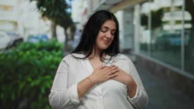 Beyaz gömlekli kadın, güneşli bir şehir caddesinde güven içinde duruyor, şehir ortamında memnuniyet ve pozitifliğini ifade ediyor, güzellik ve güveni yansıtıyor..