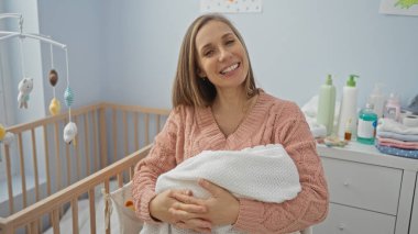 Bebek beşiği ile sıcak bir yatak odasında, bebek ürünleri ile çevrili bir şekilde gülümseyen, sükunet gösteren, annelik ve bakım ortamını besleyen bir kadın..