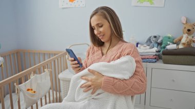Beşiğinde oyuncaklar olan rahat bir yatak odasında telefonu kullanırken kucağında bebek tutan kadın ve gülen yüz çevreyi beslerken birden fazla iş yaptığını gösteriyor.