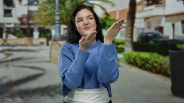 Kadın, güneşli bir cadde boyunca açık havada neşeli bir süveter giyerek neşeli şehir atmosferini sergilerken mutlu bir şekilde el kol hareketi yapıyor..