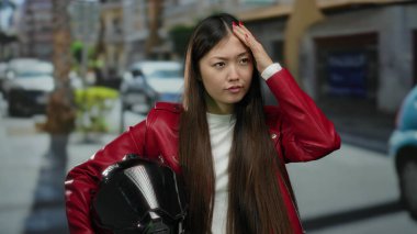 Kırmızı ceketli, siyah miğferli, şehir manzarası bulanık ve park halindeki arabalarla çevrili bir kadın. Modern yaşam tarzını çağrıştırıyor..